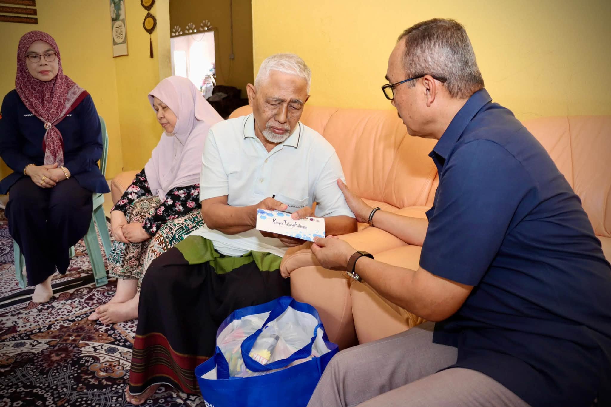 Seorang pengunjung menyerahkan sampul kepada warga emas yang duduk di sofa, dengan dua wanita berada berhampiran.
