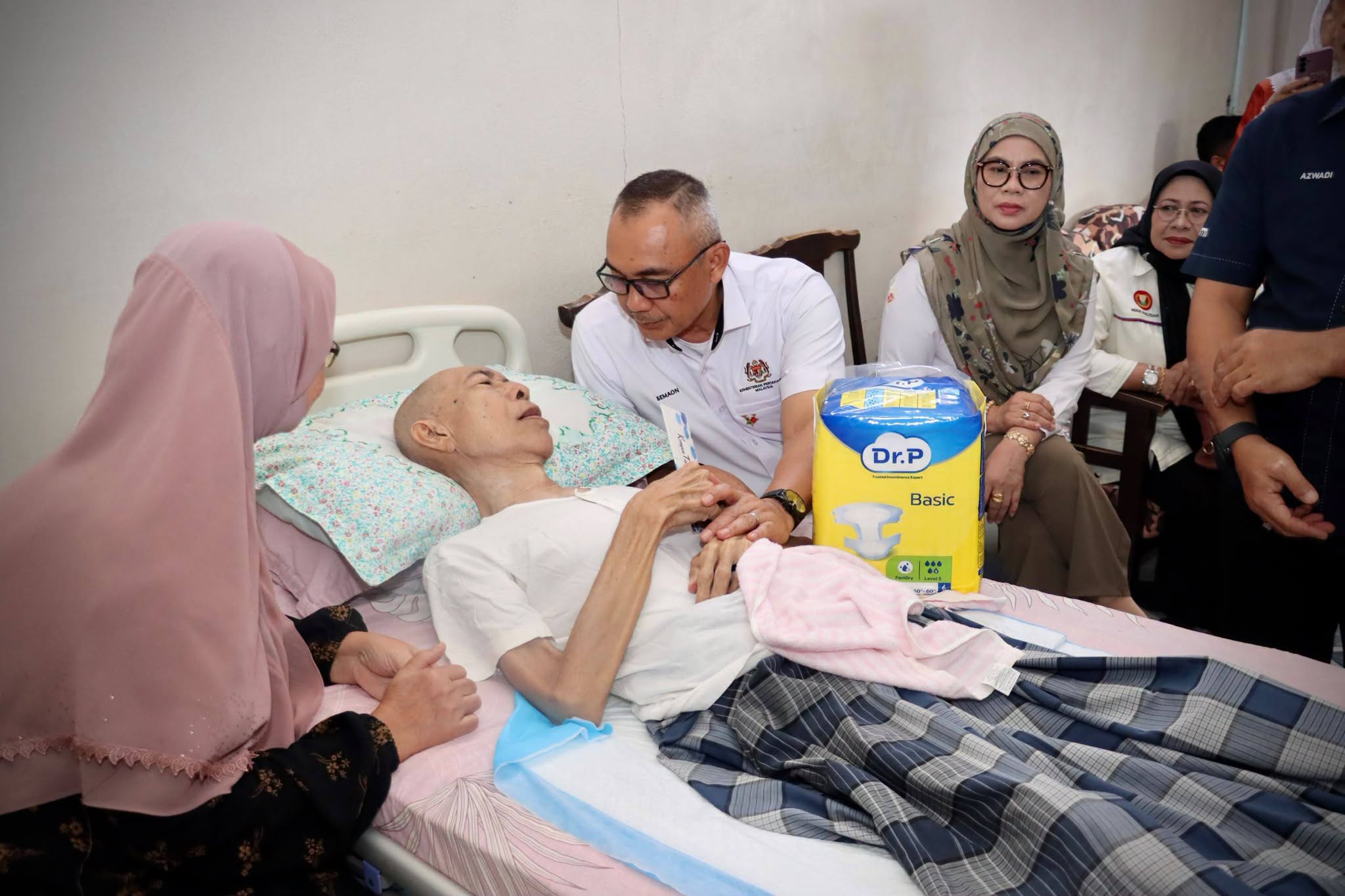 Seorang pegawai berbaju putih menziarahi pesakit yang berehat di katil bersama beberapa orang pelawat.